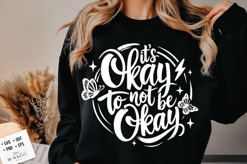 May include: Black sweatshirt with the white text "It's Okay to not be Okay" in a circular design with butterfly and star graphics. The sweatshirt is a crew neck style.