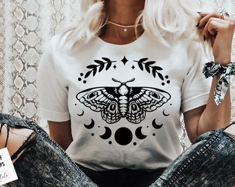 May include: A white t-shirt with a black and white moth design. The moth is surrounded by a crescent moon and stars. The design is centered on the shirt.