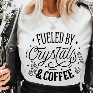 May include: A white t-shirt with black text that reads "Fueled by Crystals & Coffee". The text is in a stylized font and includes small crystal drawings.