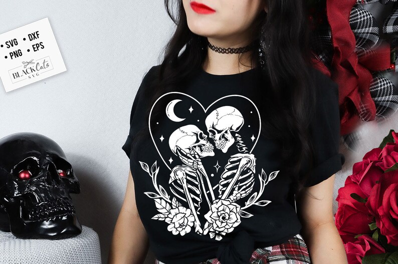 May include: Black t-shirt featuring a white graphic of two skeletons kissing within a heart shape, adorned with a crescent moon and stars. The skeletons are surrounded by flowers and leaves.