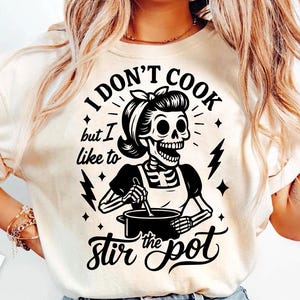 Op de afbeelding: Crèmekleurig T-shirt met een zwart-witte afbeelding van een skelet met een schort en een pot aan het roeren. De tekst luidt: "I DON'T COOK but I like to stir the pot."
