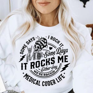 May include: White sweatshirt with a black graphic featuring a skeleton with a bun and the text "Some Days It Rocks Me, Medical Coder Life." The design includes lightning bolts and the words "We're Rockin'."