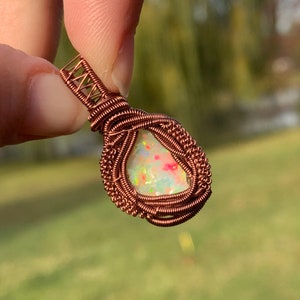 May include: A copper wire wrapped pendant featuring a white opal gemstone with iridescent colors. The pendant is held by a finger.