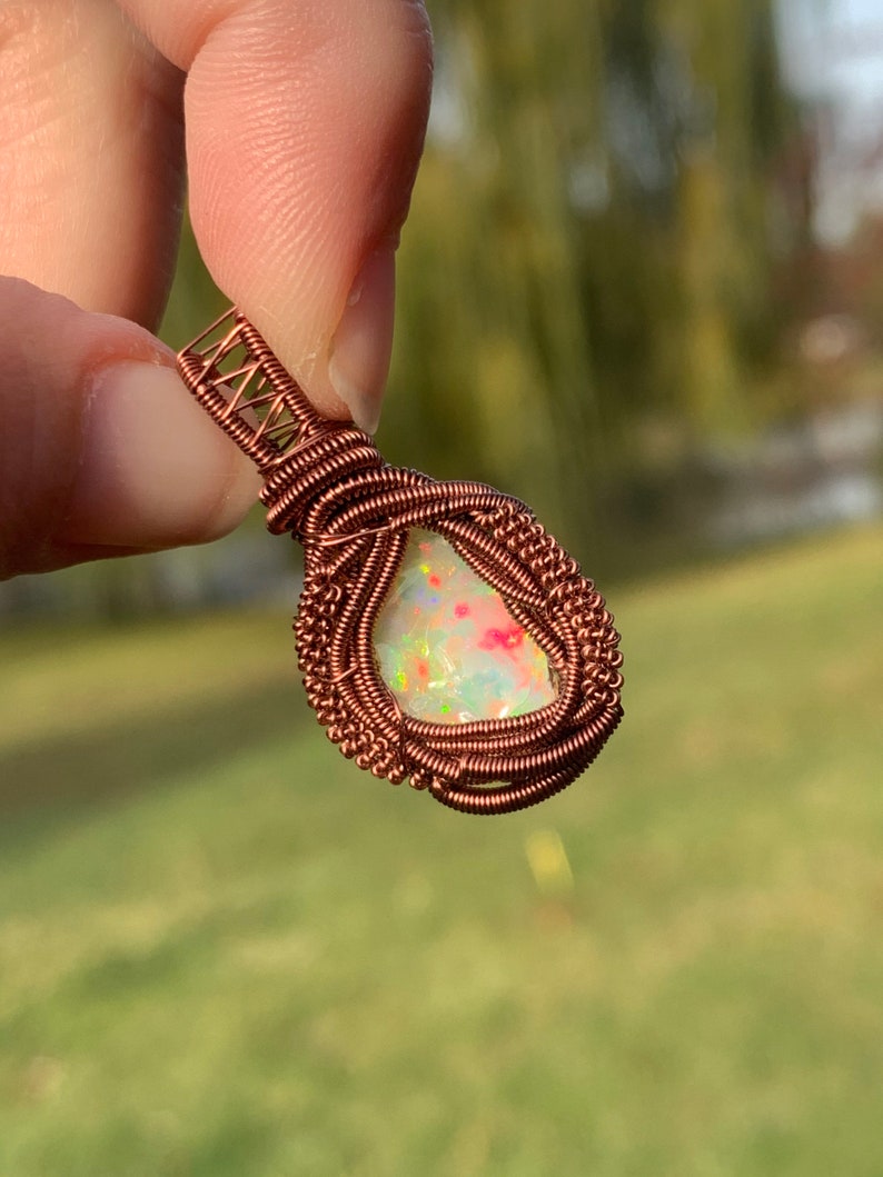 May include: A copper wire wrapped pendant featuring a white opal gemstone with iridescent colors. The pendant is held by a finger.