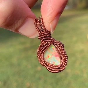 May include: A copper wire wrapped pendant featuring a white opal with pink and green inclusions. The pendant is held by a hand.