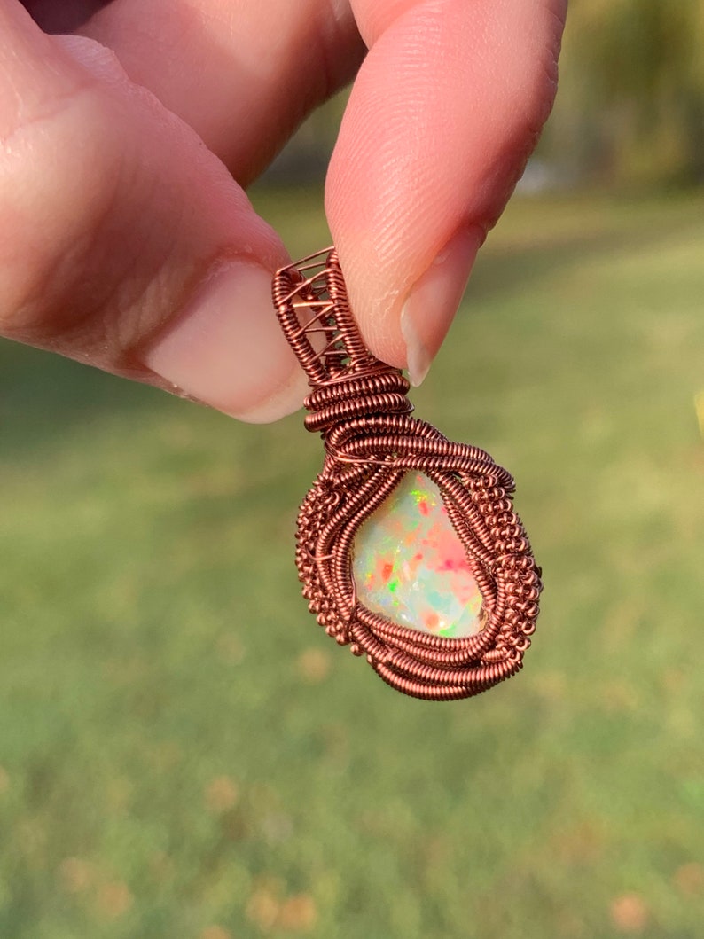 May include: A copper wire wrapped pendant featuring a white opal with pink and green inclusions. The pendant is held by a hand.