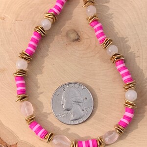 May include: A pink and white striped beaded necklace with gold accents and clear quartz stones. The necklace is laid out on a wooden surface with a US quarter for scale.