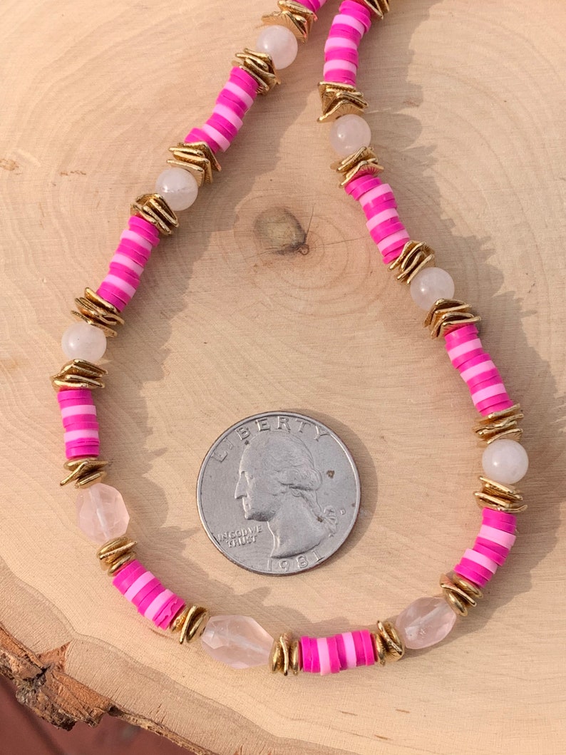 May include: A pink and white striped beaded necklace with gold accents and clear quartz stones. The necklace is laid out on a wooden surface with a US quarter for scale.