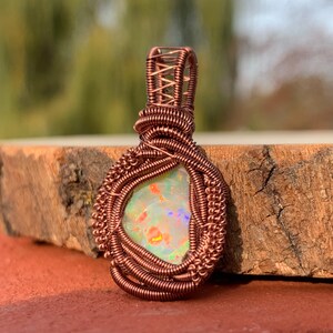 May include: A copper wire wrapped pendant featuring a white opal with iridescent rainbow colors. The pendant is on a wooden surface.