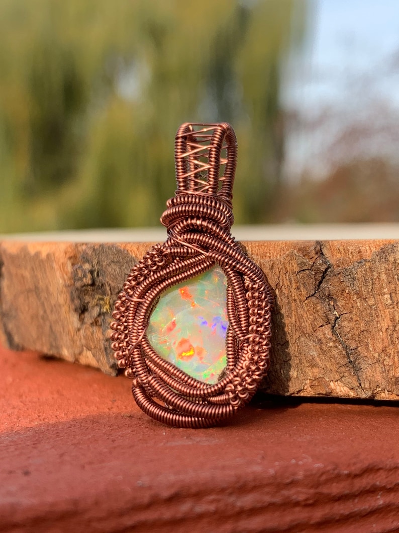 May include: A copper wire wrapped pendant featuring a white opal with iridescent rainbow colors. The pendant is on a wooden surface.