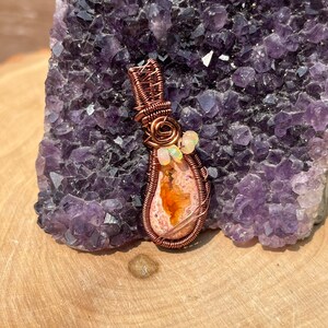 May include: A copper wire wrapped pendant featuring a teardrop-shaped orange and white opal gemstone. The pendant is accented with small white beads and is displayed on a purple amethyst cluster.