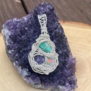 May include: A handcrafted pendant featuring a green gemstone, an opal, and small purple stones, all intricately wire-wrapped in silver. The pendant rests on a purple amethyst geode, showcasing a natural, artistic design.