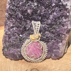Puede incluir: Un colgante de piedra rosa con un envoltorio de alambre plateado y detalles de ópalo. El colgante se muestra en un racimo de amatista púrpura.