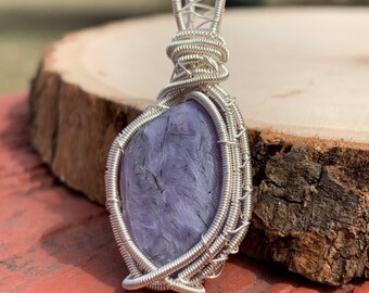 May include: A silver wire wrapped pendant featuring a purple and gray gemstone. The pendant is on a wooden surface.