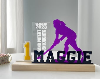 May include: A purple silhouette of a field hockey player with a stick, a black acrylic plaque with the name "MAGGIE" and the number "1" in yellow, and a clear acrylic plaque with the text "CLASS OF 2023" and "LAND PATENT GOLDEN KNIGHTS".