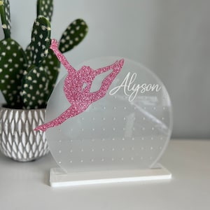 May include: A clear acrylic earring holder with a pink glitter gymnast silhouette and the name "Alyson" inscribed on the front. The holder has a white base and a circular top with multiple holes for earrings.