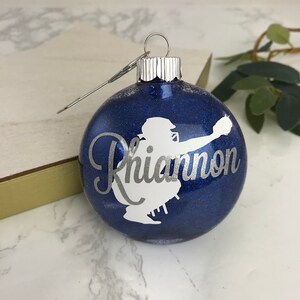 May include: A blue glitter Christmas ornament with a silver baseball catcher silhouette and the name "Rhiannon" in silver script. The ornament has a silver metal cap and hanger.