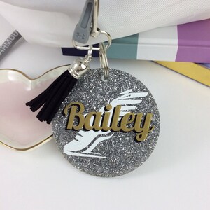 May include: A round, glittery silver keychain with the name "Bailey" in gold lettering. A white graphic of a figure skater is below the name. The keychain has a black tassel and a silver clasp.