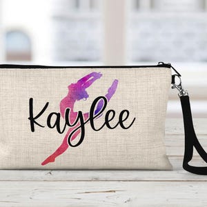 May include: A beige canvas makeup bag with a black zipper and a black strap. The bag has a watercolor image of a gymnast in a pink, purple, and blue silhouette. The name "Kaylee" is written in black cursive lettering.