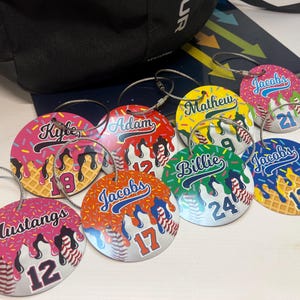 May include: A collection of personalized baseball-themed luggage tags. Each tag features a unique design with names, numbers, and a baseball graphic. The tags have a colorful, dripped ice cream cone design with sprinkles and a metal loop for attachment.