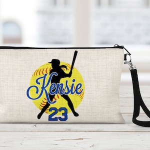 May include: A beige canvas zippered pouch with a black strap. The pouch features a yellow softball with a black silhouette of a player swinging a bat. The text "Kensie 23" is printed in blue on the softball.