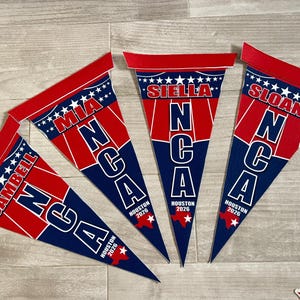 May include: Four red, white, and blue pennant flags with the text "NCA" and names like "Mia" and "Siella". Each flag has "Houston 2026" and a Texas outline. The flags are on a light wood surface.