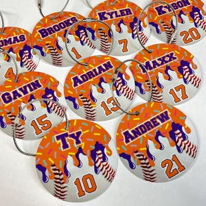 May include: A collection of baseball-themed luggage tags. Each tag is round with a white baseball design, orange frosting, and sprinkles. Names and numbers are printed on each tag, with a silver wire loop for attachment.