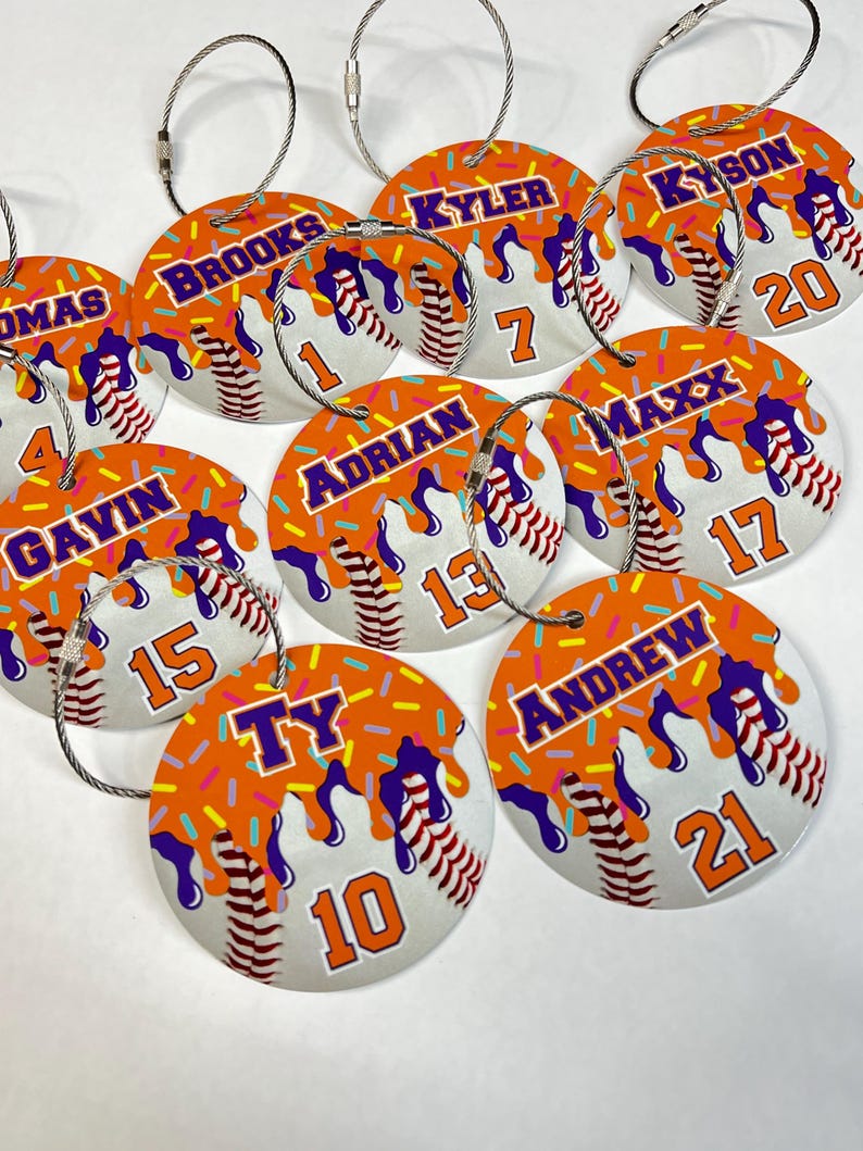 May include: A collection of baseball-themed luggage tags. Each tag is round with a white baseball design, orange frosting, and sprinkles. Names and numbers are printed on each tag, with a silver wire loop for attachment.
