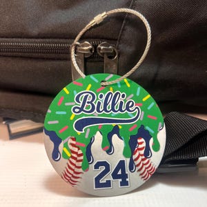 May include: A round luggage tag with a green background and the name "Billie" in white script. The tag features a baseball design with the number "24" and a silver wire loop for attachment.