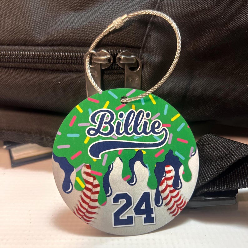 May include: A round luggage tag with a green background and the name "Billie" in white script. The tag features a baseball design with the number "24" and a silver wire loop for attachment.