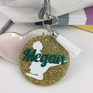 May include: Gold glitter circle keyring with a white silhouette of a person and the name Megan in green letters. The keyring has a white tassel.
