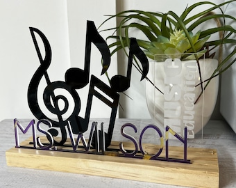 Può includere: Una targa in legno con un design di note musicali nere e il nome "MS WILSON" in lettere nere. La targa è su una base in legno e ha un cartello in acrilico trasparente con il testo "Music Education" in lettere bianche.