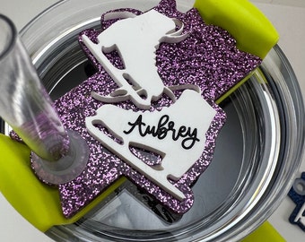 May include: A purple glitter tumbler lid with a white silhouette of a figure ice skating. The figure is holding a skate and the name "Aubrey" is written on the skate.