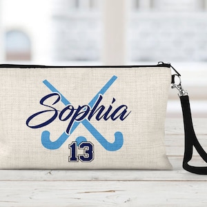 May include: A beige canvas cosmetic bag with a black zipper and a black strap. The bag has a blue and navy blue design of two crossed field hockey sticks with the name "Sophia" and the number "13" printed on it.
