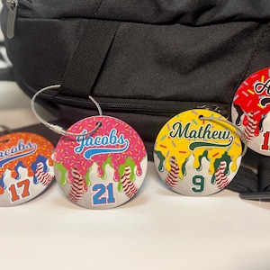 May include: Four round baseball-themed luggage tags with names and numbers. Each tag features a baseball design with a name and number, and a colorful drip design. The tags are attached to a black backpack.
