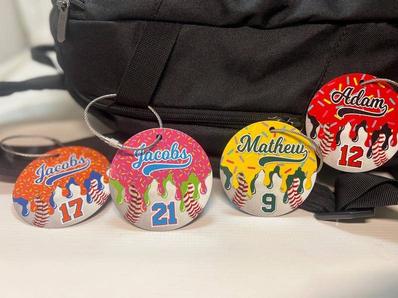 May include: Four round baseball-themed luggage tags with names and numbers. Each tag features a baseball design with a name and number, and a colorful drip design. The tags are attached to a black backpack.