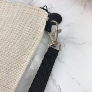 May include: A close-up of a beige canvas bag with a black strap and a silver clasp. The strap is attached to the bag with a black metal ring.