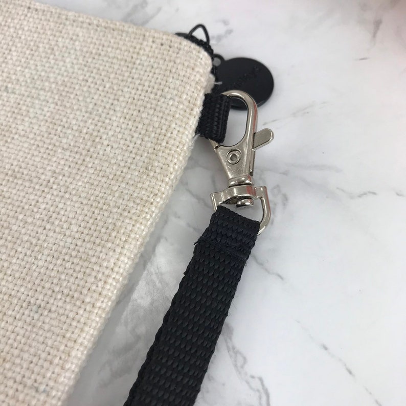 May include: A close-up of a beige canvas bag with a black strap and a silver clasp. The strap is attached to the bag with a black metal ring.