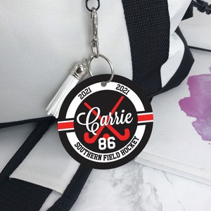May include: A black and white round keychain with a red and white stripe. It says "2021 Carrie 86 2021 Southern Field Hockey" in white text.