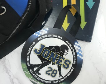 May include: A round, green and black football-themed personalized name tag with the text "PANTHERS FOOTBALL 2019" and the name "JONES" with the number "28" in gold.