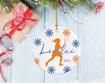 May include: White ceramic Christmas ornament with a blue and orange lacrosse player silhouette, snowflakes, and the name "Lorelei" and the year "2022".