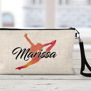 May include: A beige canvas zippered pouch with a black zipper and a black strap. The pouch has a silhouette of a gymnast in orange and red with the name "Marissa" printed in black.