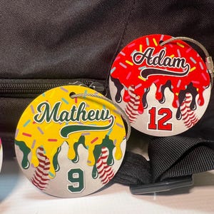 May include: Two round luggage tags with a baseball theme. One tag is yellow with the name "Mathew" and the number "9". The other tag is red with the name "Adam" and the number "12". Both tags have a silver wire loop.