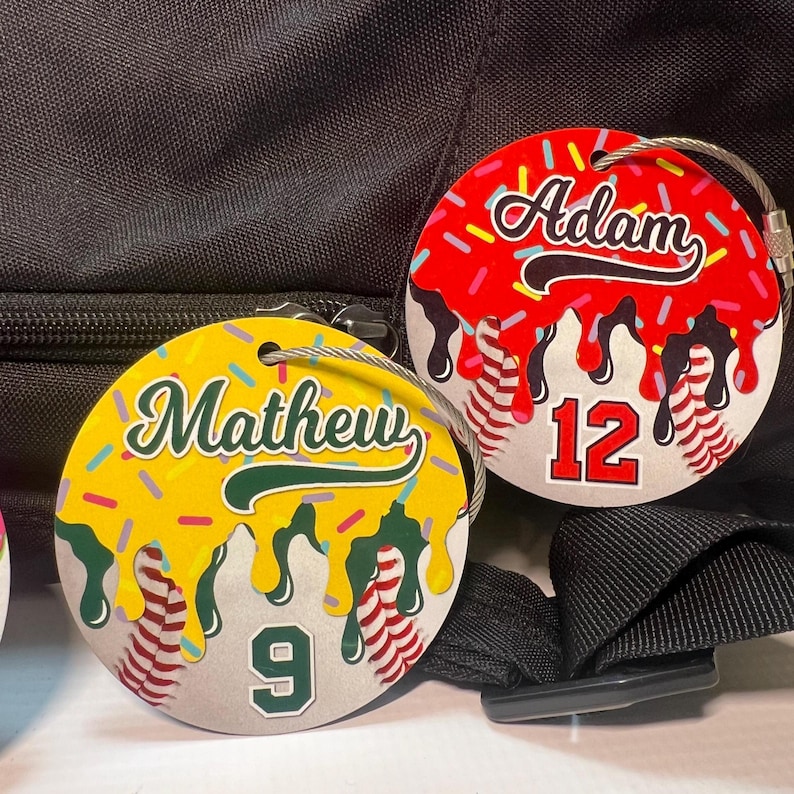 May include: Two round luggage tags with a baseball theme. One tag is yellow with the name "Mathew" and the number "9". The other tag is red with the name "Adam" and the number "12". Both tags have a silver wire loop.