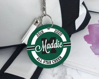 May include: Green and white round keychain with the text "2019" and "2020" on either side of a black letter "M" with the name "Maddie" written in cursive above it. The text "All Star Cheer" is written below the letter "M".