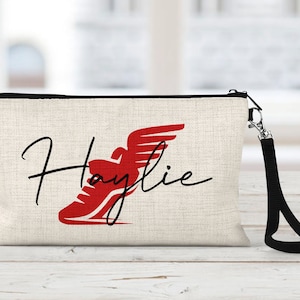 May include: A beige canvas makeup bag with a black zipper and a black strap. The bag has a red running shoe graphic and the name "Haylie" written in black cursive.