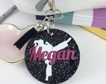 May include: Black and white glitter circle keychain with a white silhouette of a person doing a karate kick. The name "Megan" is written in pink glitter letters.