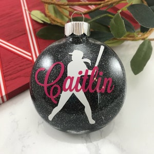 May include: A black glitter Christmas ornament with a white silhouette of a softball player holding a bat. The name "Caitlin" is written in pink script across the ornament. The ornament has a silver cap.