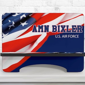 May include: A blue and red personalised phone stand with an American flag design. The stand has a shelf and the text "AMN BIXLER U.S. AIR FORCE" is printed on the front.