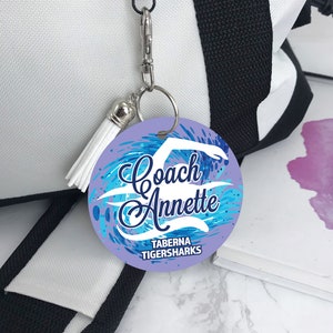 May include: A purple and blue round keychain with a white silhouette of a swimmer and the text "Coach Annette Taberna Tigersharks".